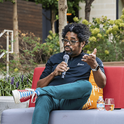 jason perry a person with a dark blue shirt and teal pants sits holding a microphone on an outdoor couch